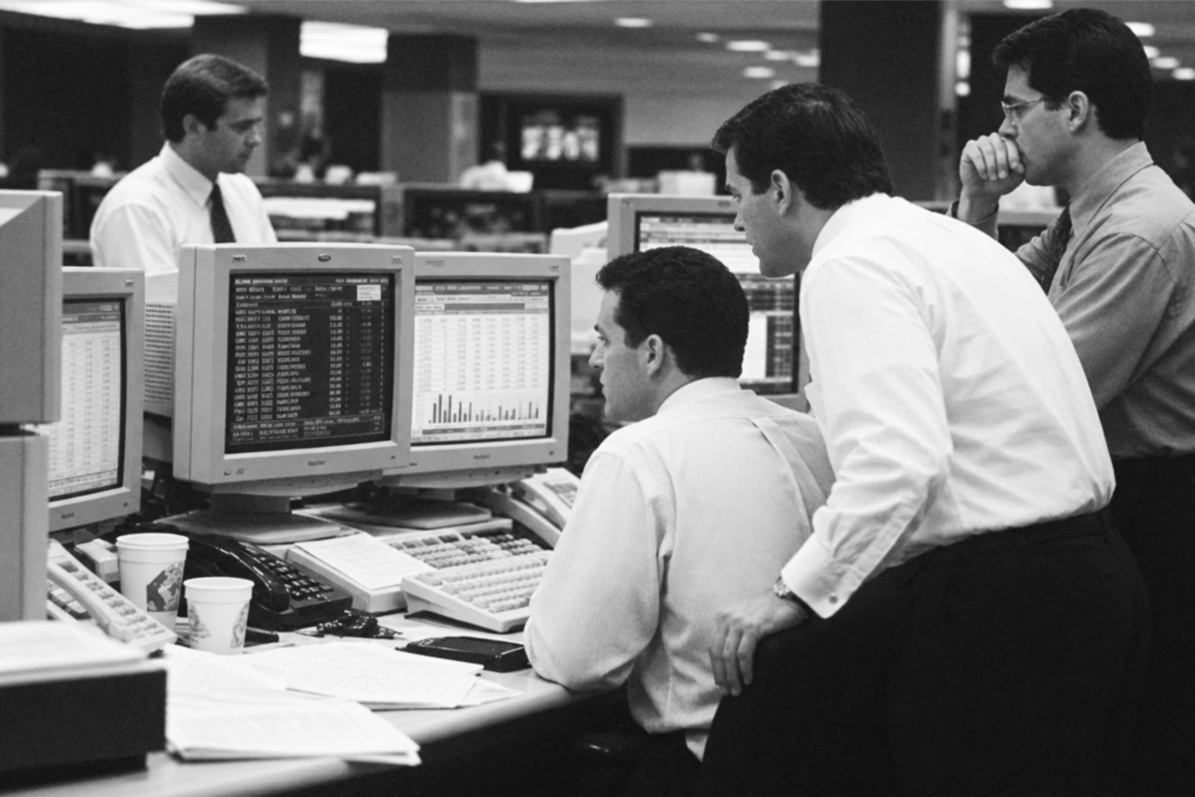 Traders in shirts and ties at a desk of CRT monitors, two of them leaning over a colleague's shoulder to read his screen.